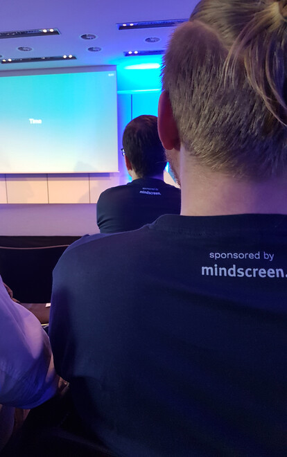 Zwei Teilnehmer der Neos Konferenz mit Konferenz-T-Shirts auf denen das Logo des T-Shirt-Sponsors mindscreen gedruckt ist.